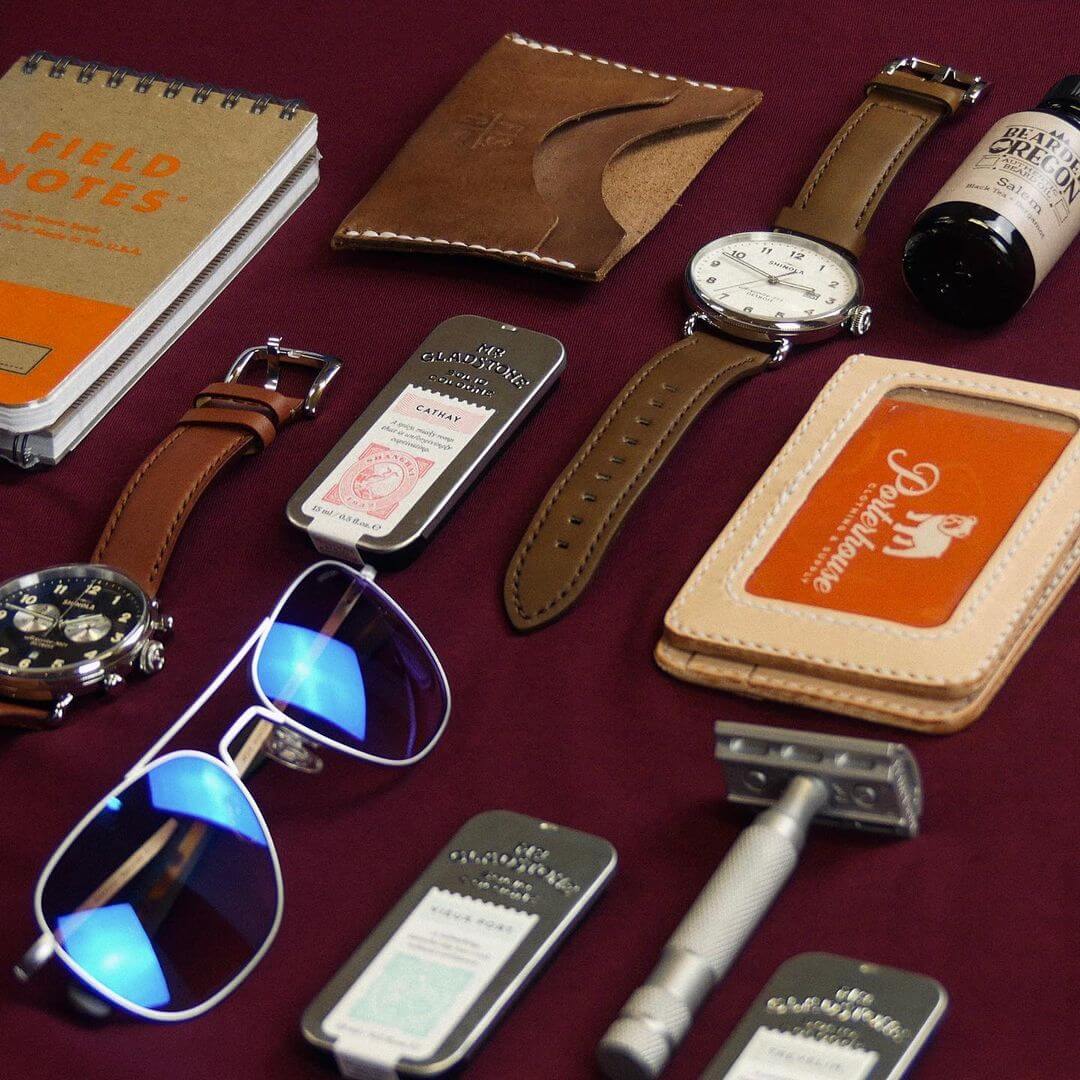 Display of men's accessories laid out, like cologne, sunglasses, watches, Field Notes and leather goods