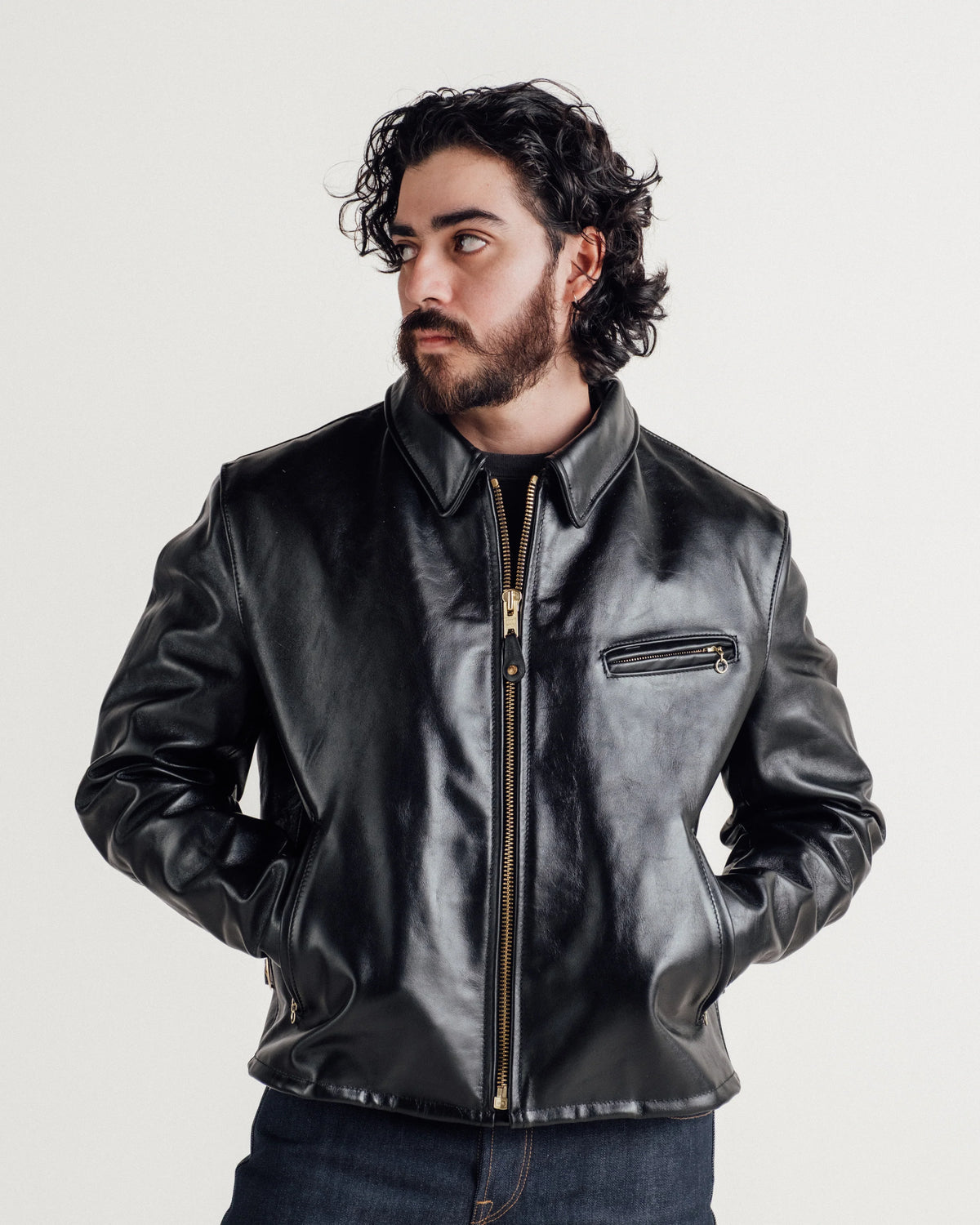 Male model wearing black horsehide leather jacket with denim, hands in jacket pockets, looking off-camera left. See product details for more information.