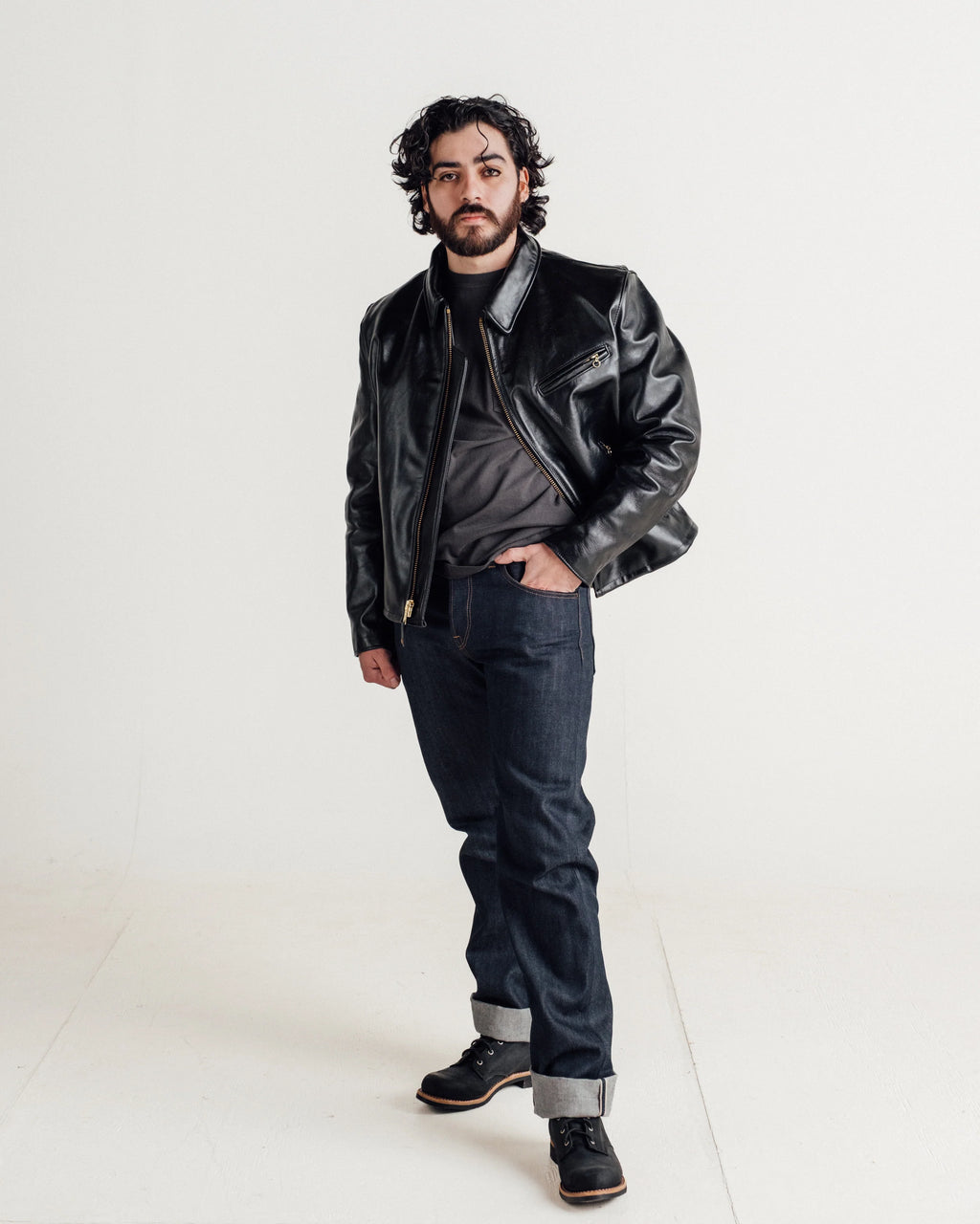Bearded male model with dark curly shoulder-length hair sporting black horsehide leather jacket, styled with selvedge denim and black boots. One hand in pants pocket. See product details for more information.