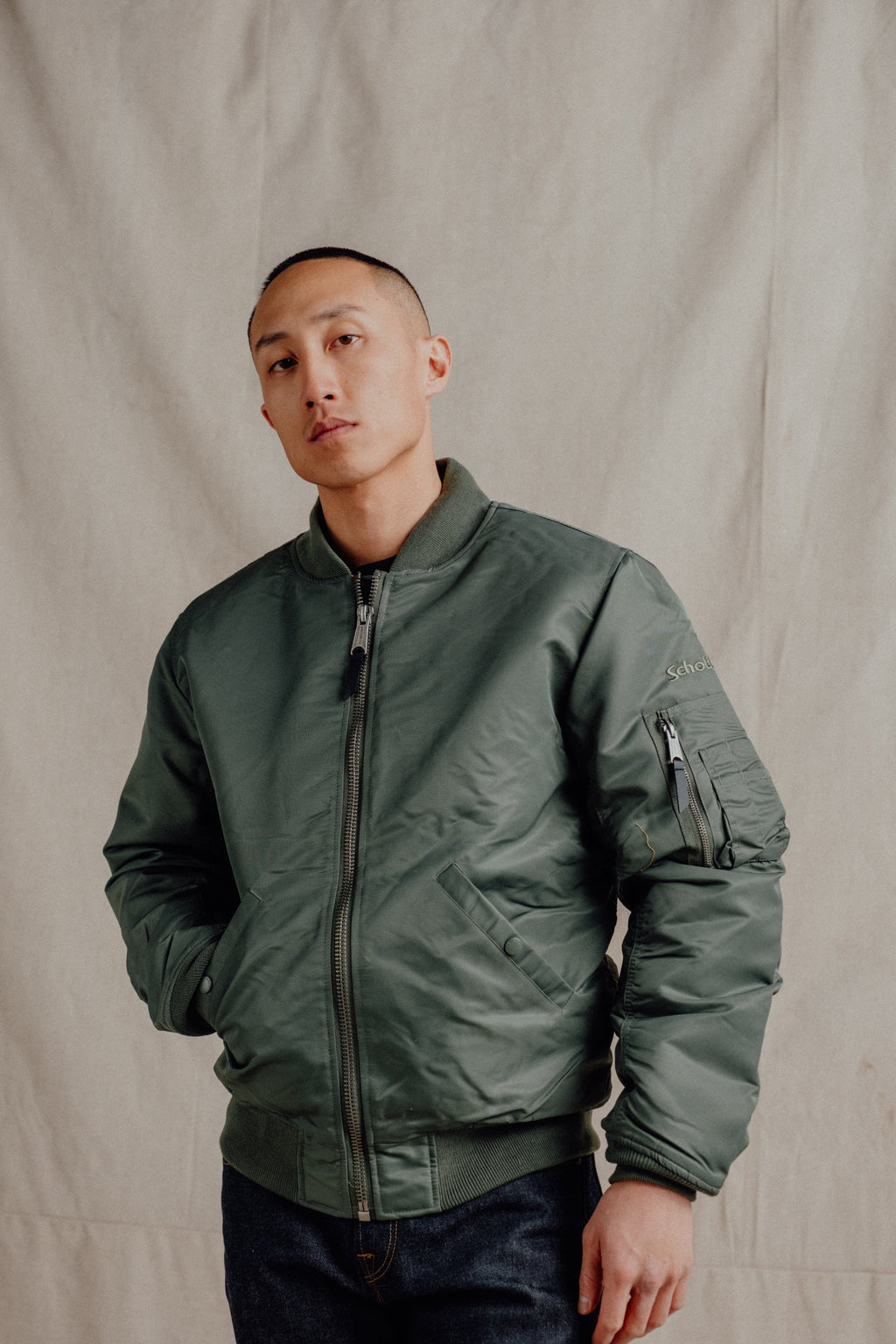 Male model wearing sage bomber jacket. One hand in hand warmer pocket. Beige fabric backdrop. See product details for more information.