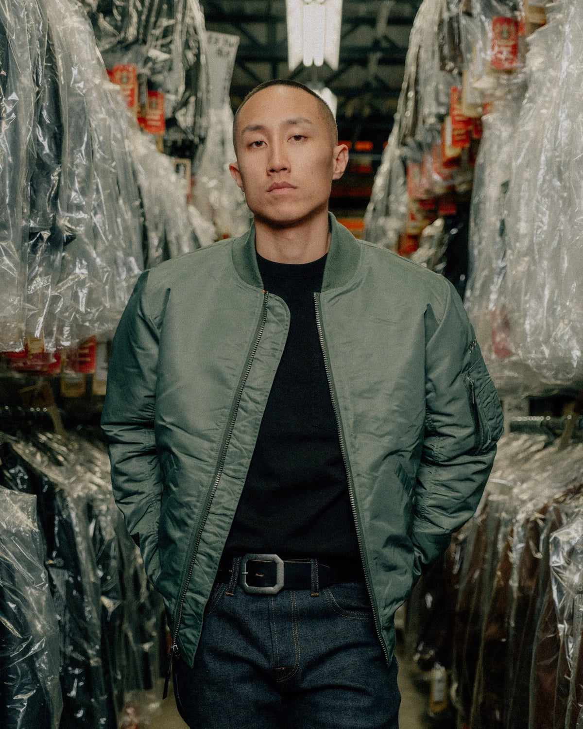 Male model sporting sage bomber jacket in a factory with two-tier rows of plastic-covered jackets on either side of him. See product details for more information.