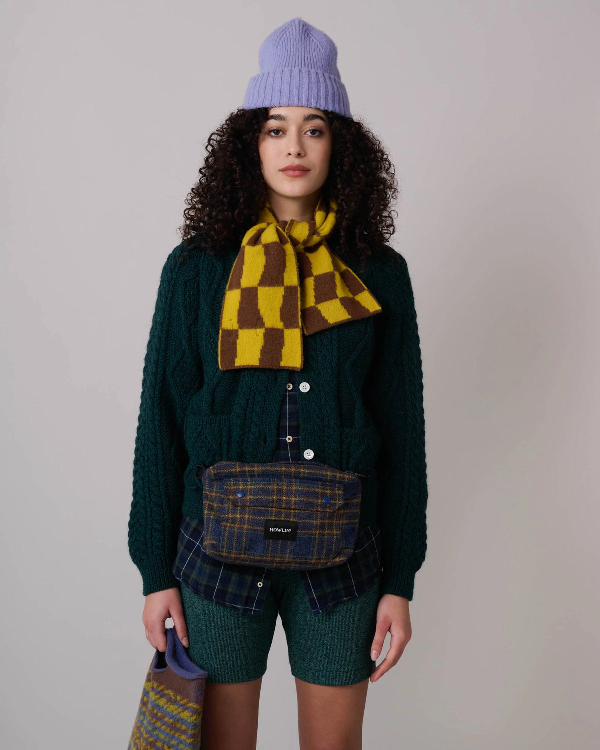 Female model wearing lavender wool watch cap with cuff over frizzy dark hair. Also sporting a yellow and brown checked scarf, forest green cable knit cardigan over blackwatch check button down and elastic forest green shorts. A blue and brown check shoulder bag hangs in front of her and a geometric print yellow and brown wool handbag is at her side. See product details for more information.