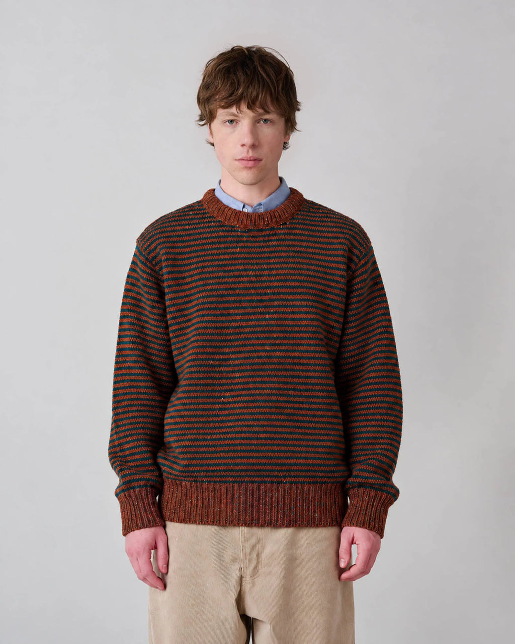 Young man wearing Broken Finger Dream in Cinnamon—black striped crewneck sweater on brown field—over a solid blue button-down and khaki corduroy trousers. See product description for more details.