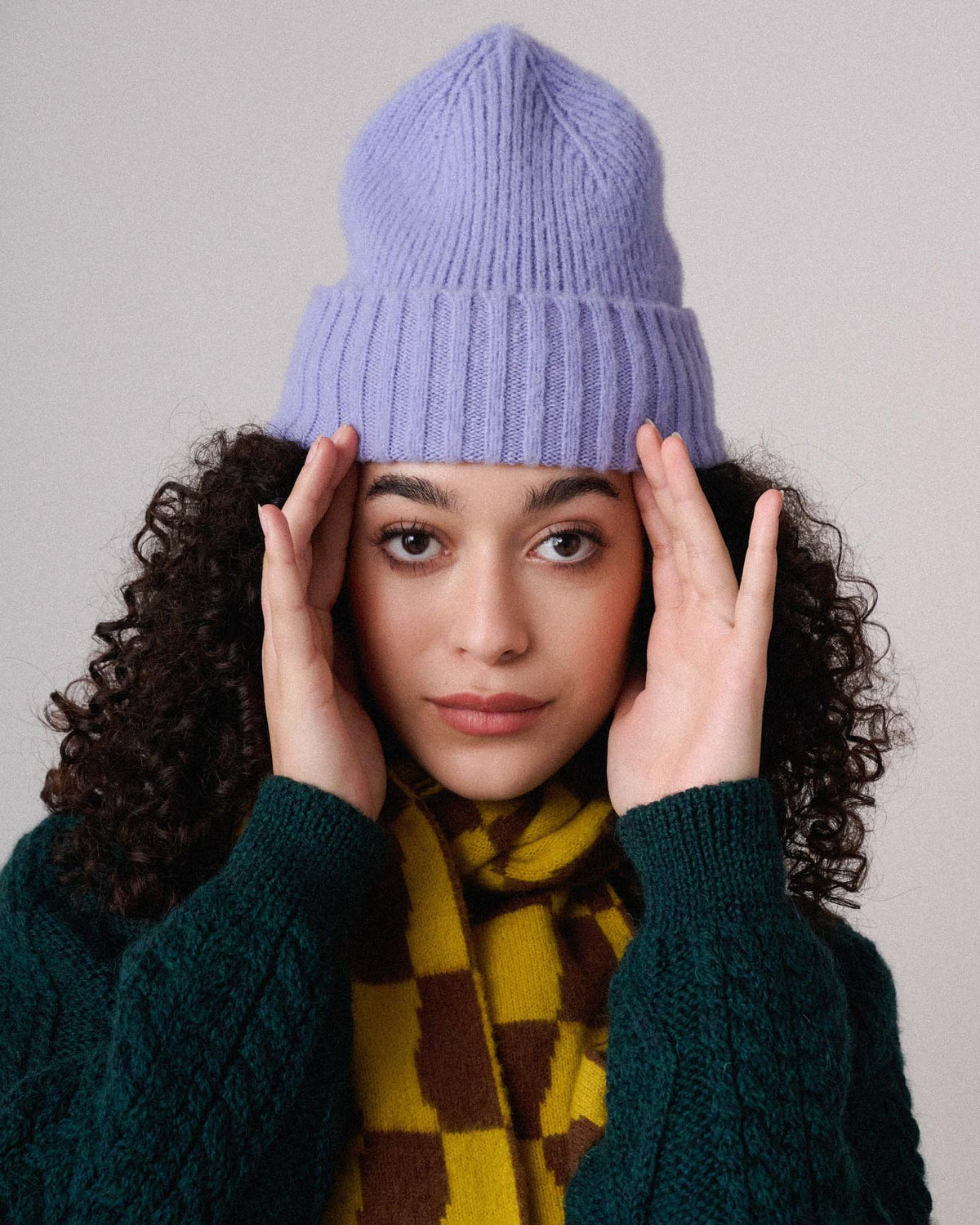 Headshot of female model wearing lavender wool watch cap with cuff over frizzy hair. Yellow and brown check scarf around her neck, forest green cable knit cardigan on. Holding her hands to the sides of her face as though she's looking through a porthole. See product details for more information.